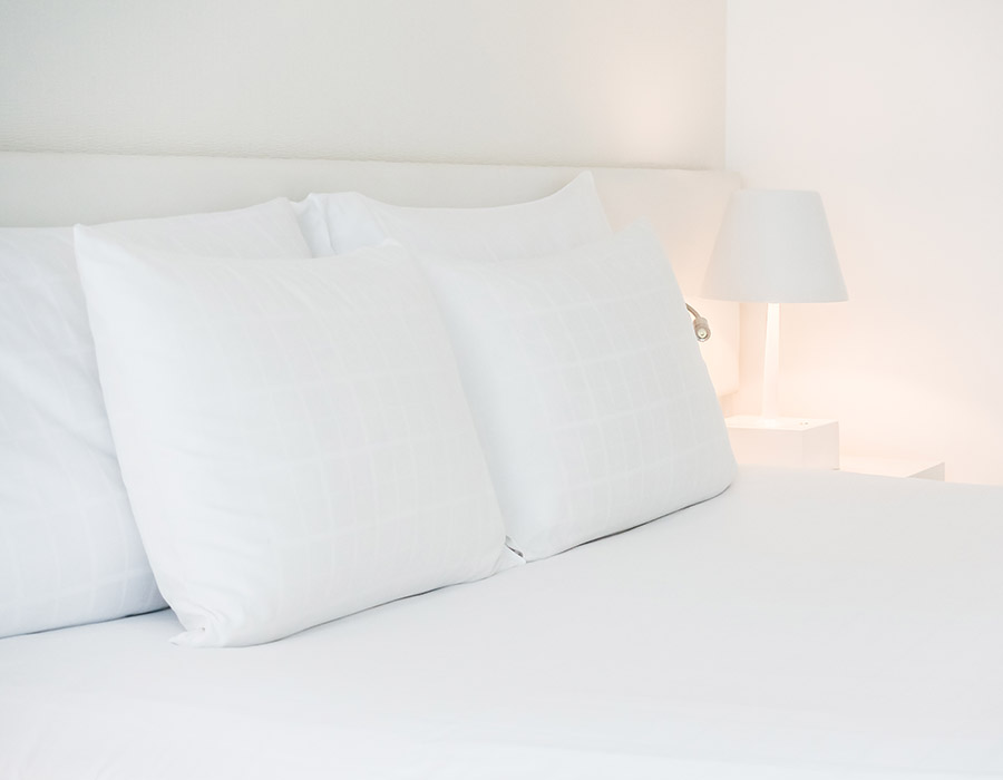 Cama de hotel. Primer plano almohadas, cojines y sábanas en color blanco. La mesita, lámpara y paredes también son de color blanco.