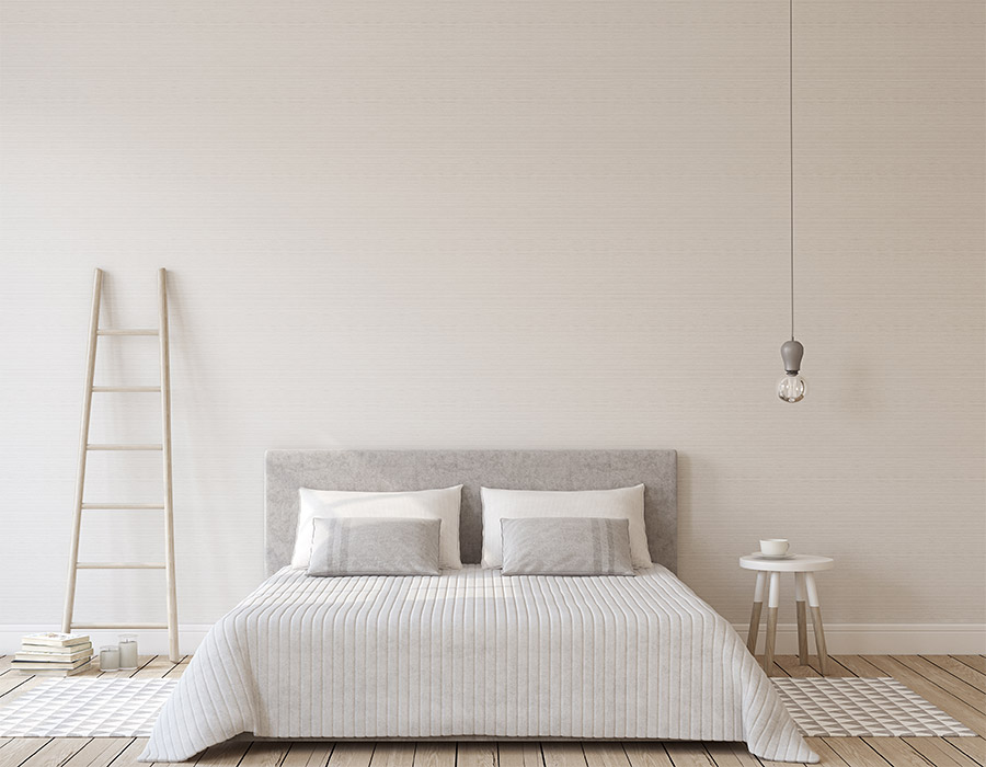 Dormitorio blanco. Dormitorio estilo nórdico. Vista frontal cama con ropa de cama en tonos grises muy claros con almohadones en el mismo tono y blancos. Mesita de noche a un lado, alfombras en tonos marrones a ambos lados de la cama y suelo parquet de madera. 